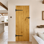 Lifestyle A tall, narrow wooden door with vertical planks in a warm honey finish, styled with black metal barn-style hardware. The door is set within a white wall in a bright, modern farmhouse-style living space featuring a sofa, wooden ceiling beams, and a kitchen in the background.